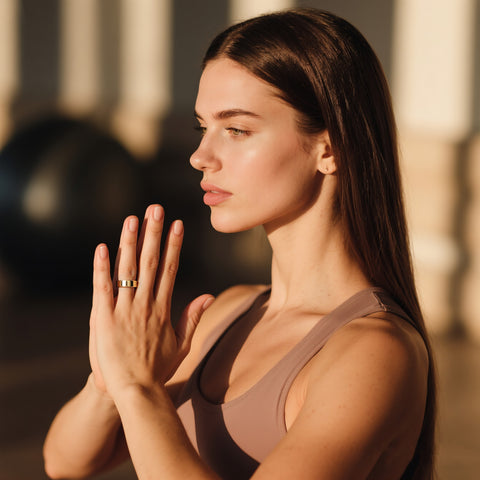 Side-profile woman practicing yoga in warm sunlight, hands in prayer pose, wearing a gold POBOVi smart ring for wellness tracking.